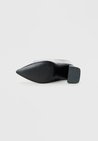 Zapato de tacón negro con puntera afilada, con parte superior de cuero liso y suela texturizada, que presenta un tacón de goma grueso para mayor estabilidad.