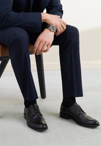 Brązowe skórzane buty garniturowe z detalami brogue, w połączeniu z czarnymi skarpetkami i granatowymi spodniami, umieszczone na jasnym dywanie. Żadne inne elementy nie są widoczne.