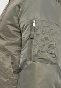 Olive roheline paks jakk, millel on tõmblukuga tasku vasakul varrukal ja nähtavad õmblusdetailid. Sile nailonmaterjal, struktureeritud disain.