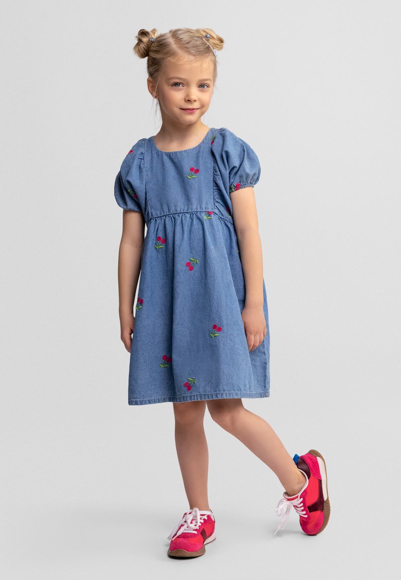 Jeune fille aux cheveux blonds coiffés en deux chignons, portant une robe bleue brodée de cerises et des baskets roses, debout avec une jambe légèrement levée.