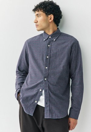 Young man with curly hair wearing a navy and red checkered button-up shirt, white undershirt, and dark pants, looking to the side.