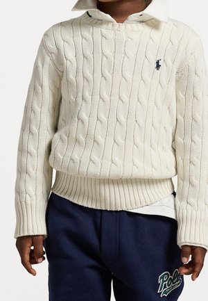 Kind draagt een witte kabelgebreide trui over een wit overhemd met kraag en donkerblauwe joggingbroek met een groen Polo-logo.