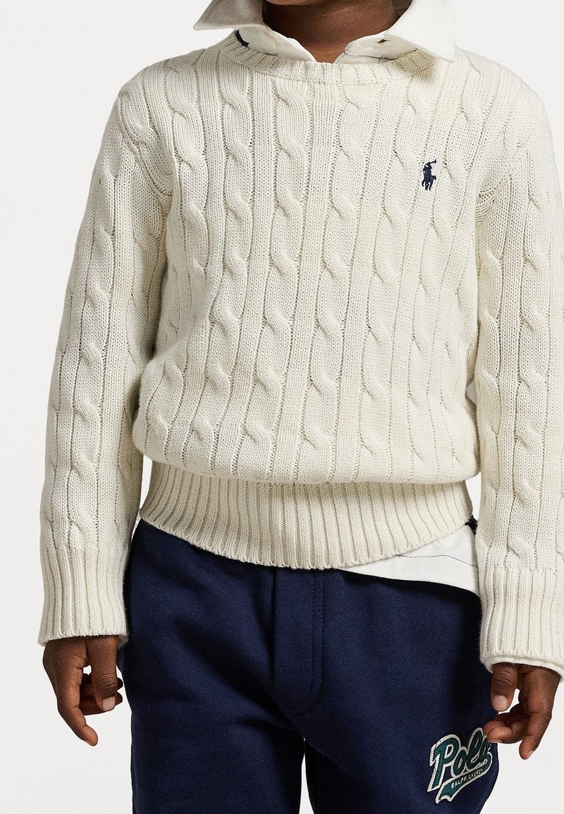 Enfant portant un pull en maille torsadée blanc par-dessus une chemise blanche avec col et un pantalon de survêtement bleu marine avec un logo Polo vert.
