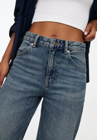 Siniset korkeavyötäröiset farkut, joissa on klassinen viiden taskun muotoilu, kontrastivärinen ompelu ja napitus, sekä haalistunut, tekstuuri pinta.