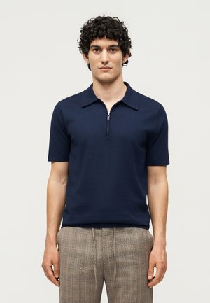 Mand med krøllet mørkt hår iført navyblå kortærmet skjorte med lynlås og beige ternede bukser, stående mod en lys ensfarvet baggrund.