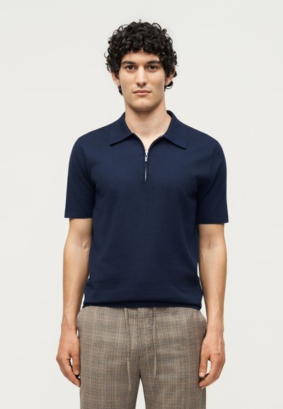 Homme aux cheveux bouclés et foncés portant une chemise bleu marine à manches courtes avec fermeture éclair et un pantalon beige à carreaux, debout devant un fond clair uni.