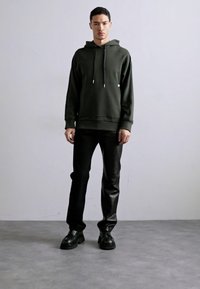 Olivenfarbener Kapuzenpulli mit verstellbaren Zugbändern, gerippten Bündchen, kombiniert mit schwarzen Lederhosen und schwarzen klobigen Schuhen.