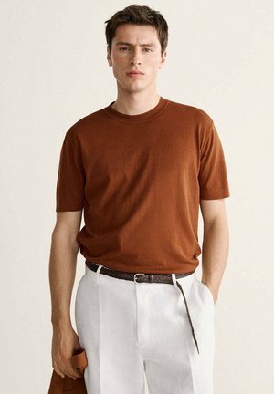 Giovane uomo che indossa una camicia marrone a maniche corte infilata in pantaloni bianchi con una cintura marrone intrecciata, che tiene un tessuto abbinato in una mano.