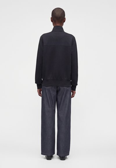 Personne debout de dos, portant une veste noire et un jean bleu foncé ample sur un fond clair uni.