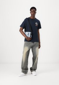 Camiseta marina con estampado gráfico, bolso bandolera azul claro, jeans baggy de lavado claro y zapatillas blancas, de pie contra un fondo neutro.