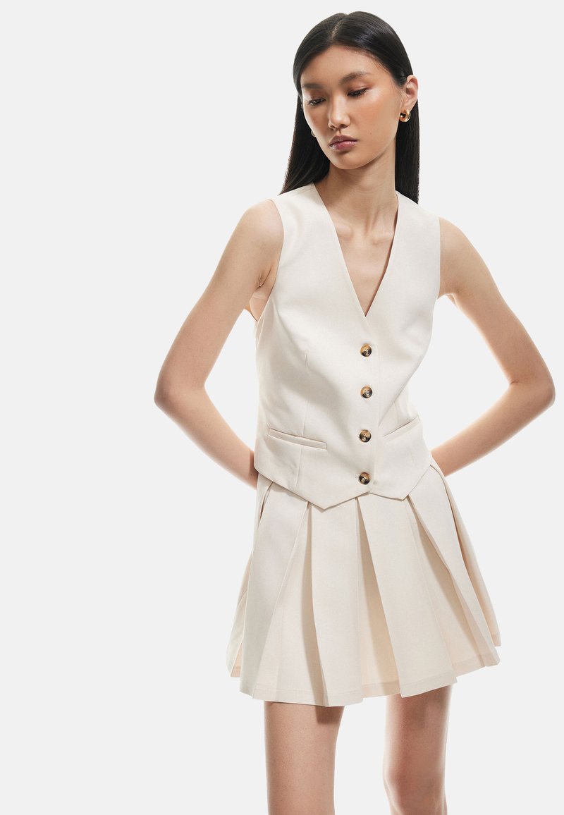 Gilet senza maniche color crema con scollatura a V profonda, cinque bottoni dorati e gonna mini plissettata. Tessuto liscio con forma strutturata e tasche laterali.