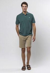 Tumši zaļa polo kreklā ar apkakli, bēšas bikses šortos un brūnas sandales. Krekls rotāts ar mazu logo uz krūtīm.