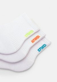 Tri páry bielych ponožiek s rebrovanou štruktúrou. Každý pár má na manžete farebný neónový logo "STWD." v zelenej, oranžovej a modrej farbe.