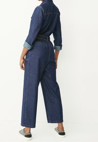 Combinaison en denim bleu foncé avec des manches longues, des poignets retroussés, une taille ceinturée et des jambes larges. Comprend deux poches arrière et des coutures visibles.