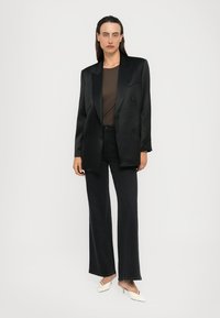 Vrouw in een zwarte dubbelzijdige blazer, een bruine top, zwarte broek en witte lage hakken, staande tegen een simpele witte achtergrond.