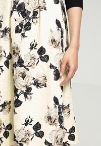 Gonna midi floreale in crema con motivi di rose nere e beige. Tessuto texturizzato con pieghe, con una mano appoggiata sulla gonna.