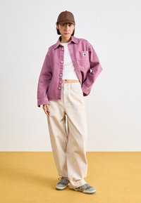 Kvinne som har på seg brun caps, oversized lilla jakke, hvit crop top, vide beige bukser og grå joggesko, stående på gul gulv mot en hvit vegg.