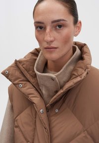 Braune Steppweste mit hohem Kragen, diagonal gesteppt, mit Druckknöpfen und sichtbaren Nähten über einer beigen Strickschicht darunter.