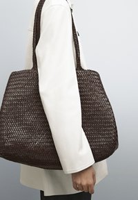 Brown, woven tote bag with a rectangular shape and two long handles, resting against a white coat, with a smooth texture and braided pattern.