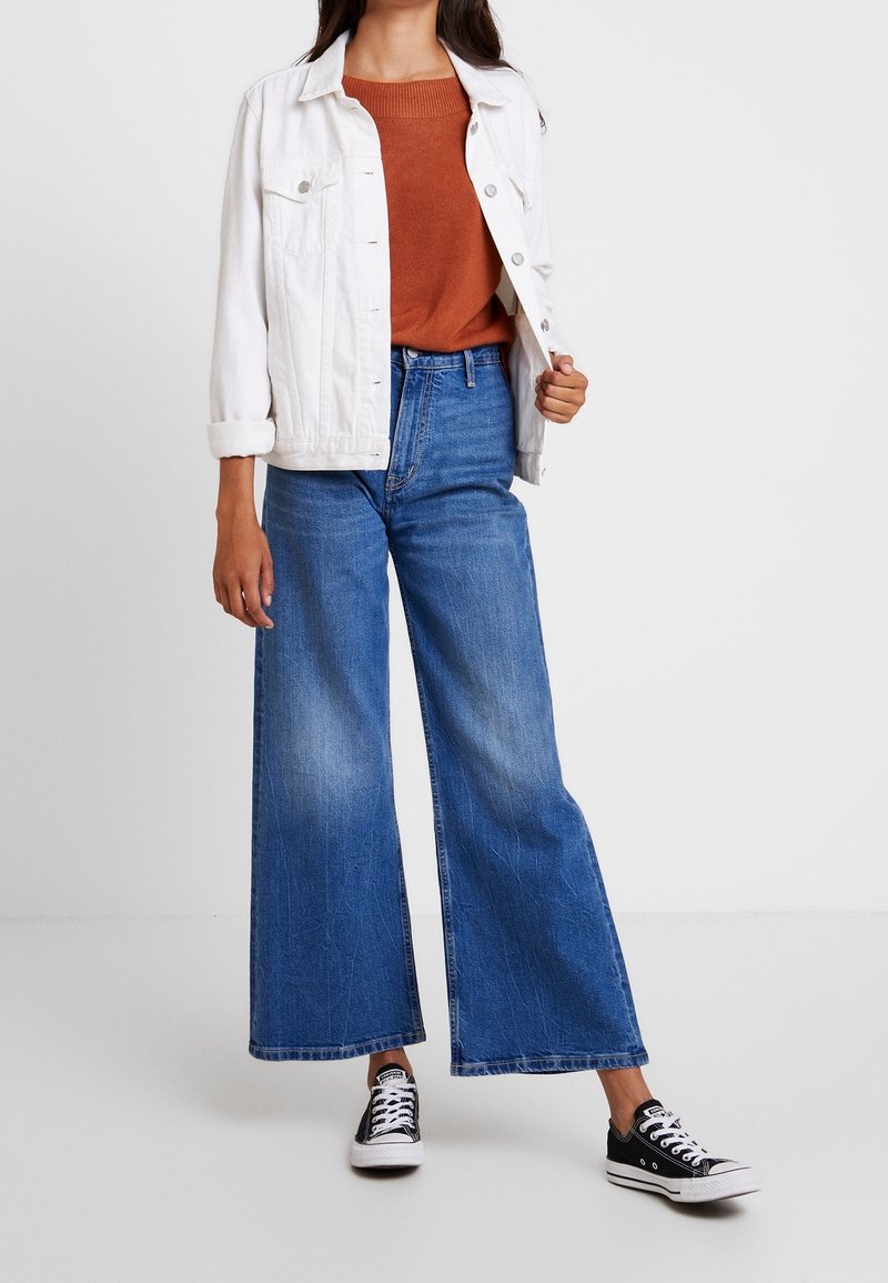 Femme portant une veste en jean blanche, un pull couleur rouille, un jean bleu à jambes larges et des baskets noires basses, debout devant un fond uni.