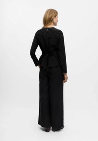 Haut noir à manches longues avec un lien à la taille et un détail en trou de clé au dos, associé à un pantalon large noir et des bottines. Texture lisse.