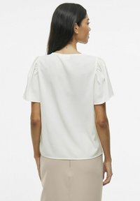 Blusa bianca a maniche corte con scollatura quadrata sul dietro e dettagli arricciati sulle spalle. Tessuto morbido con vestibilità comoda.
