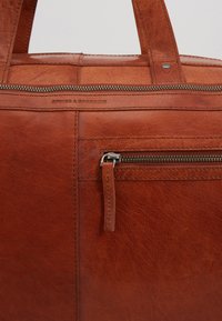 Sac en cuir marron avec une finition texturée et une poche zippée. L'affichage inclut des détails de couture et un logo de marque subtilement embossé.