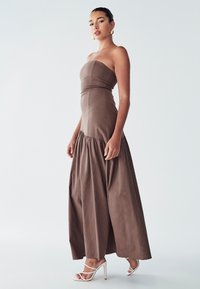 Braunes trägerloses Maxikleid mit tailliertem Oberteil und gerafftem Rock, das eine glatte Textur aufweist. Kombiniert mit weißen Absatzsandalen.