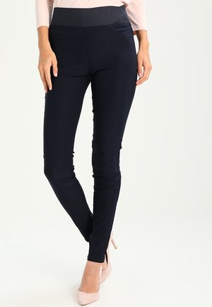 Woman wearing fitted dark navy pants with high waist and light pink heels, standing with legs crossed against plain background.