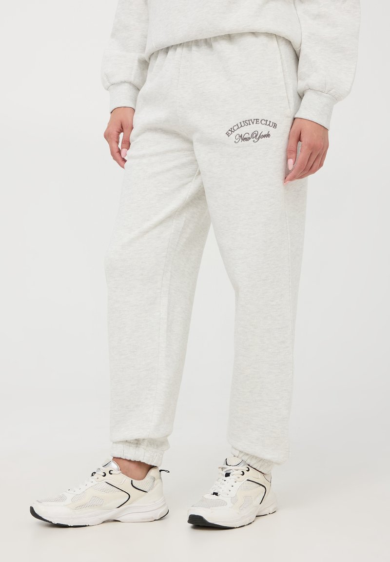 Pantaloni da tuta in cotone grigio chiaro con vita e polsini elasticizzati. Presentano la scritta "EXCLUSIVE CLUB New York" ricamata in marrone sulla coscia sinistra.