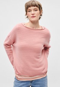 Camisola rosa de malha com gola ampla em formato barco, mangas compridas e bainha canelada. Por baixo, uma camisa de manga comprida às riscas em cores suaves.