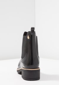 Bottes Chelsea noires brillantes avec des panneaux latéraux élastiques, une languette de traction et une semelle en caoutchouc à motif. Équipées d'un petit accent doré sur le côté.