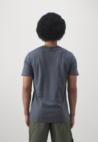 Camiseta de manga corta en gris oscuro para hombre con cuello redondo y detalle del logo en la parte posterior, fabricada en un tejido suave y transpirable. Combinada con pantalones cortos verdes.