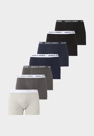 Lihaseline mees seisab paljajalu, kandes tumedaid bokserpükse valge vöökohaga, millel on kiri Jack & Jones, vaadates vasakule tavalise tausta ees.
