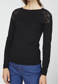 Musta neulepusero pyöreällä pääntiellä, pitkillä hihoilla ja pitsikoristelulla olkapäillä; valmistettu sileäpintaisesta neuloksesta.