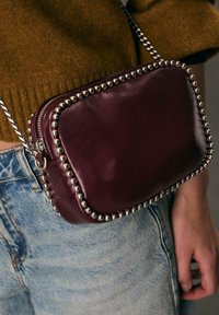 Bordeaux læder crossbody taske med sølvmetal nagledekorationer, rektangulær form, lynlås lukning og en aftagelig kæde skulderrem.