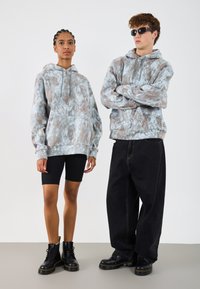 Dva modely nosiace zladené mikiny s mramorovým vzorom v svetlomodrej a hnedej farbe, v kombinácii s čiernymi šortkami a veľkými čiernymi nohavicami.