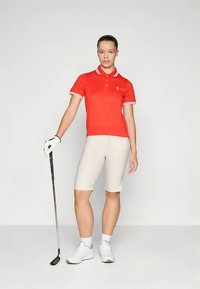 Κόκκινο πόλο πουκάμισο με λευκό γιακά, μπεζ σορτς και λευκά αθλητικά παπούτσια. Κλαμπ γκολφ στο αριστερό χέρι, σε όρθια στάση, ουδέτερο φόντο.