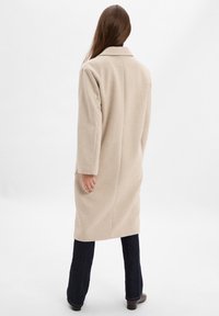 Beige Wollmischungsmantel mit strukturiertem Design, langen Ärmeln und Kragen. Der Stoff hat eine glatte Textur und wird mit dunkelblauen, ausgeweiteten Jeans kombiniert.