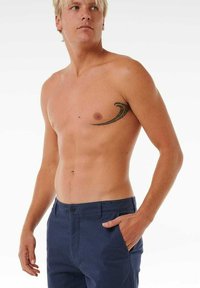 Chino blu navy con una texture liscia, dotati di chiusura a bottone e tasche laterali, indossati da una persona senza maglietta con un tatuaggio.