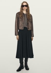 Žena s dlouhými vlnitými vlasy, oblečená v tmavých slunečních brýlích, hnědé kožené bundě, šedém topu, černé plisované midi sukni a černých vysokých kozačkách.