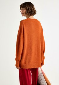 Donna con i capelli castani corti che indossa un maglione arancione oversize e pantaloni rossi, mentre tiene in mano una coperta piegata multicolore su uno sfondo bianco.