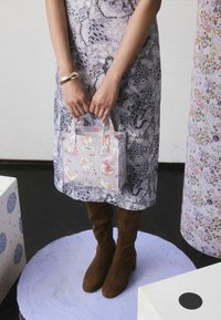Blumenmuster, helllila Handtasche mit kurzen Griffen. Enthält Illustrationen von Katzen und Hunden. Getragen zu einem gemusterten Kleid und braunen Stiefeln.