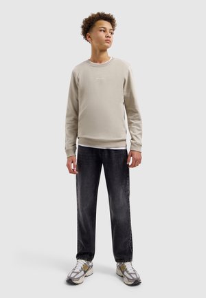 Tienerjongen staand, gekleed in een beige trui, zwarte spijkerbroek en witte sneakers, kijkt iets zijwaarts tegen een lichte achtergrond.