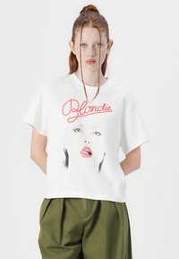 Bijela pamučna majica s grafičkim otiskom ženske lice s crvenim usnama i natpisom "Blondie" u crvenom rukopisnom fontu. Kratki rukavi, opušteni kroj.