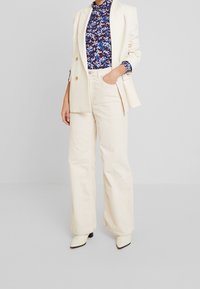 Beige kordfløjl brede bukser parret med en marineblå blomstret højhalset bluse med lange ærmer og en cremet blazer med guldtonede knapper.