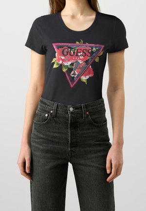 Femme portant un t-shirt noir ajusté avec un logo Guess floral rose et un jean gris foncé taille haute sur fond uni.