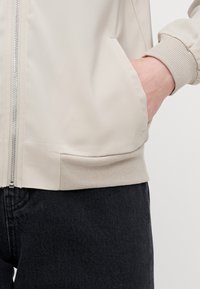 Světle béžová bomber bunda s hladkým povrchem, žebrovanými manžetami a boční kapsou; má stříbrný zip a strukturovaný detail na spodním lemu.