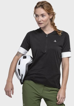 Frau mit roten Haaren im Zopf, trägt ein schwarz-weißes Radtrikot und grüne Hose, hält einen weißen Fahrradhelm unter dem Arm.