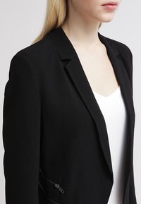 Blazer structuré noir avec revers à encoche, poches latérales zippées et finition lisse. Porté sur un haut blanc, mettant en valeur sa coupe ajustée.